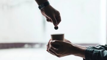 يد تضع عملة في كوب ورقي، يدي شخصين متقابلتين في مشهد يعكس مفهوم التبادل والدعم.