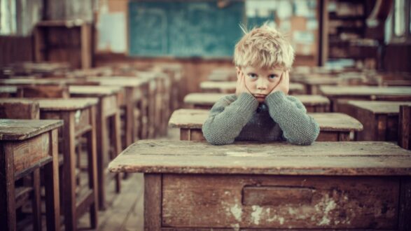 طفل يجلس وحيدًا في فصل دراسي قديم، يضع يديه على خديه، ويبدو عليه الملل أو القلق. المقاعد والطاولات الخشبية تعطي إحساسًا بالزمن الماضي، مع جدران فصول المدرسة التقليدية.