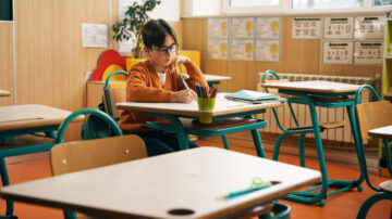 طفل يجلس بمفرده في فصل دراسي، يكتب في دفتره مع وجود أدوات مكتبية ملونة حوله. تظهر المقاعد والطاولات الخشبية في الخلفية، مع جدران مزينة بمعلومات تعليمية.