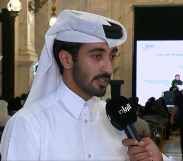 رجل يرتدي الزي التقليدي القطري يتحدث إلى مايكروفون خلال حدث رسمي، مع وجود جمهور في الخلفية.