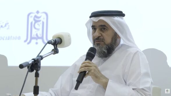 رجل يرتدي الزي التقليدي العربي يتحدث في حدث رسمي، مع وجود ميكروفون أمامه. تظهر خلفه شعارات مؤسسية على شاشة.