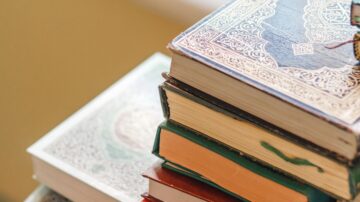 Stack of ornate, colorful books with decorative spines, resting on a surface with a blurred background. The books feature intricate designs and varying colors, showcasing a mix of textures and styles.