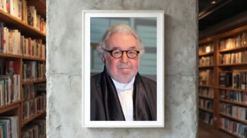 صورة لرجل مسن يرتدي نظارات وقميصًا أبيض مع سترة داكنة، يظهر بابتسامة ودودة. الصورة مؤطرة ومعلقة على جدار رمادي داخل مكتبة تحتوي على رفوف مليئة بالكتب.