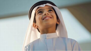 طفل إماراتي مبتسم يرتدي الكندورة والغترة، ينظر إلى الأعلى بتفاؤل. تعكس الإضاءة الساطعة في الخلفية أجواء السعادة والبراءة.