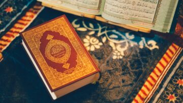 كتاب القرآن الكريم مفتوح على سجادة مزخرفة، مع نسخة مغلفة باللون الأحمر والذهبي بجانبه.
