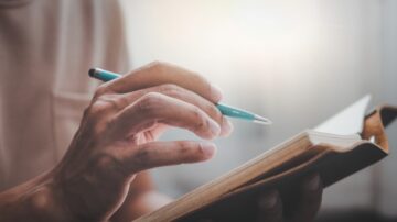 يد تشير إلى كتاب مفتوح مع قلم أزرق. تبرز الإضاءة الخلفية جوًا من التركيز والتأمل.