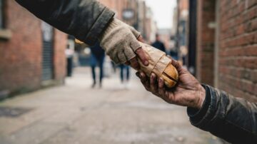 يد تمتد لتسليم طعام لشخص محتاج في شارع حضري، مع خلفية غير واضحة لأشخاص آخرين في المشهد.