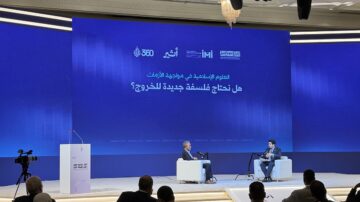 نقاش في مؤتمر حول الإعلام في عصر التكنولوجيا الحديثة، يضم شخصين يجلسان على منصة أمام شاشة تعرض سؤالاً حول الحاجة لفلسفة جديدة في الإعلام. الحضور يتابعون الجلسة في قاعة مؤتمرات.