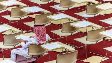 طالب يرتدي الزي السعودي يجلس وحده في قاعة امتحانات، محاطاً بمقاعد فارغة وأوراق اختبار على الطاولات.