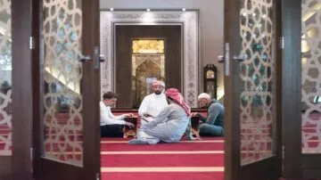 رجلان يرتديان الأزياء التقليدية يجلسان على سجادة حمراء داخل مسجد، مع شخصين آخرين يجلسان على الأرض. تظهر الأبواب الخشبية المفتوحة في المقدمة، مما يبرز الأجواء الروحية والتقليدية للمكان.