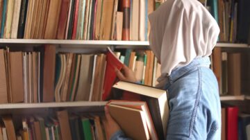 امرأة ترتدي حجابًا أبيض، تحمل مجموعة من الكتب، تتصفح رفوف المكتبة المليئة بالكتب. تعكس الصورة حب القراءة والتعلم.