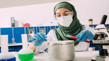 امرأة ترتدي الحجاب والنظارات الطبية وقناع الوجه في مختبر علمي، تقوم بإجراء تجربة باستخدام أنابيب اختبار تحتوي على سوائل ملونة. تظهر معدات مختبرية وأدوات علمية في الخلفية، مما يبرز بيئة البحث العلمي.
