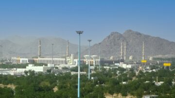 منظر بانورامي لمدينة مكة المكرمة، يظهر فيه الكعبة الشريفة والمباني المحيطة بها. تهيمن الجبال على الخلفية، وتحيط بالأشجار الخضراء. السماء صافية، مما يبرز جمال المعمار الإسلامي.