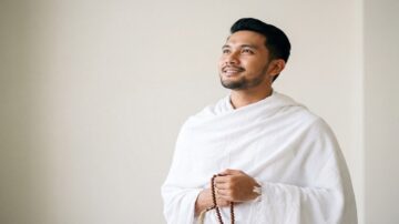 رجل يرتدي ثوبًا أبيضًا، يحمل سبحة، وينظر إلى السماء بابتسامة. تعكس الصورة لحظة تأمل وروحانية، مع خلفية بسيطة.