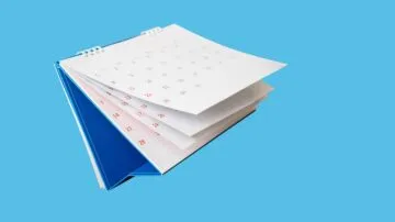 تقويم شهري مفتوح يظهر صفحات بيضاء مع تواريخ بالأحمر، على خلفية زرقاء. يوضح الترتيب والتنظيم الزمني.