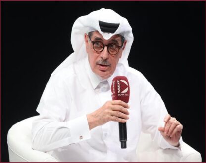رجل يرتدي الزي القطري التقليدي، بما في ذلك القميص الأبيض والغترة، يجلس على كرسي ويستخدم ميكروفونًا للحديث. يعبر عن أفكاره في حدث ثقافي أو مناقشة عامة.