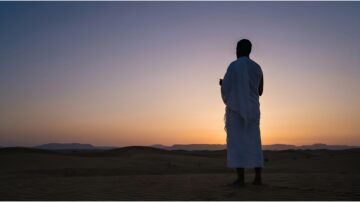 Silhouette of a man in Ihram attire standing in a desert at sunset, with soft, colorful skies and distant mountains in the background.