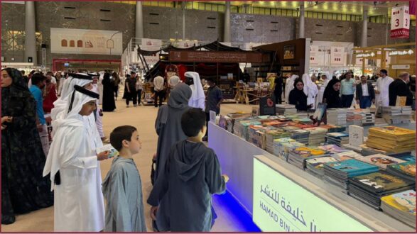 معرض كتاب في مركز ثقافي مزدحم، يظهر زوارًا يرتدون الملابس التقليدية، يتصفحون مجموعة من الكتب المتنوعة المعروضة على طاولة. في الخلفية، توجد أجنحة عرض وديكورات تقليدية، مما يضيف جوًا ثقافيًا للحدث.