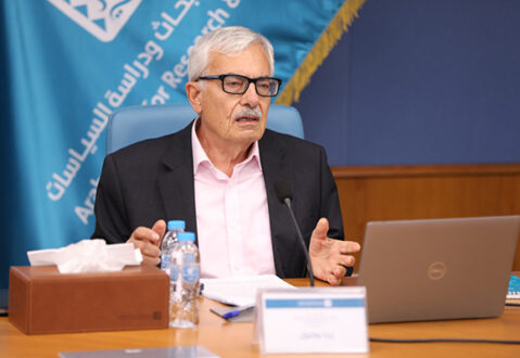 رجل مسن يرتدي نظارات وقميص وردي يتحدث في مؤتمر عبر الإنترنت، يجلس أمام جهاز كمبيوتر محمول. خلفه خلفية زرقاء تحمل شعار مؤسسة بحثية. على الطاولة زجاجة ماء وعلبة مناديل.