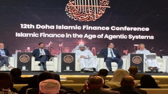 صورة من المؤتمر الثاني عشر للمالية الإسلامية في الدوحة، حيث يجلس مجموعة من المتحدثين على منصة أمام جمهور. يتناول المؤتمر موضوع المالية الإسلامية في عصر الأنظمة الوكيلة. تظهر خلفية الشاشة شعار المؤتمر مع تفاصيل حول الحدث.