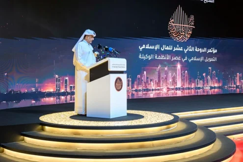 رجل يتحدث على منصة في مؤتمر إسلامي حول التمويل الإسلامي، مع خلفية تظهر skyline لمدينة حديثة، وأضواء موجهة نحو المنصة.