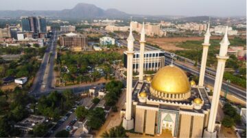 Aerial view of the National Mosque in Abuja, Nigeria, showcasing its golden dome and towering minarets, surrounded by urban buildings and greenery, with hills in the background.