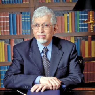 رجل مسن يرتدي بدلة داكنة وقميصًا أزرق، يجلس أمام مكتبة تحتوي على كتب ملونة. يتسم بتعابير وجه ودودة، مع نظارات وشعر رمادي.