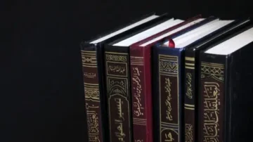 Une collection de livres reliés, présentant des couvertures en cuir noir et rouge, avec des titres dorés en arabe. Les livres sont alignés sur une étagère sombre, mettant en avant leur élégance et leur richesse culturelle.