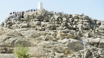 Vue du mont Arafat, avec des pèlerins rassemblés sur les rochers, symbole important du Hajj en Arabie Saoudite. Le paysage aride met en évidence la structure blanche au sommet, entourée de formations rocheuses.