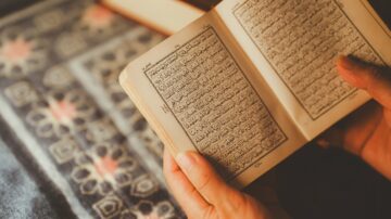 كتاب مفتوح يتضمن نصوص باللغة العربية، يتم قراءته بأيدٍ بشرية، مع وجود سجادة مزخرفة في الخلفية.