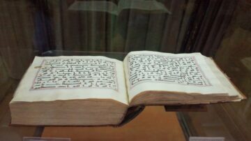 كتاب قديم مفتوح يعرض نصوص باللغة العربية، مع خطوط مزخرفة وألوان تقليدية، محفوظ داخل زجاج.