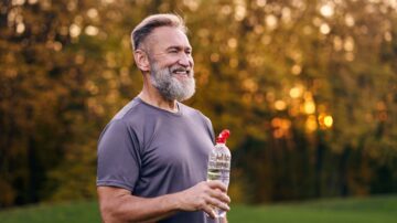 رجل مسن مبتسم يحمل زجاجة ماء، يرتدي قميص رياضي، ويقف في حديقة أثناء غروب الشمس، مع أشجار خضراء في الخلفية.