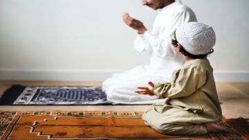 رجل يؤدي الصلاة بينما يجلس طفل صغير بجانبه على سجادة صلاة. يظهر التركيز والسكينة في الأجواء.
