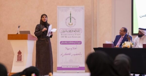 امرأة ترتدي ملابس تقليدية تتحدث في مؤتمر، أمام منصة تحمل شعار الاتحاد الدولي لعلماء المسلمين. يوجد رجال جالسون في الخلف، ومن بينهم شخص يرتدي بدلة. الطاولة مزينة بالزهور.