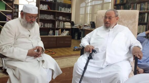 صورة لرجلين يجلسان في مكتبة. الرجل الأيسر يرتدي ثوبًا أبيض وعمامة بيضاء، بينما الرجل الأيمن يرتدي ثوبًا أبيض أيضًا ويستخدم عصا للمشي. الخلفية تحتوي على رفوف مليئة بالكتب.
