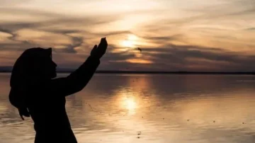 Silhouette of a person with a headscarf standing by a lake at sunset, hands raised towards the sky, reflecting warm hues on the water's surface.
