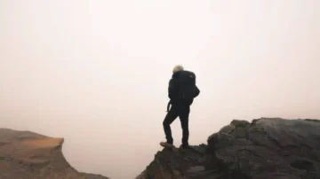 Homme silhouette sur un sommet rocheux, portant un sac à dos, face à un paysage brumeux et indéfini.