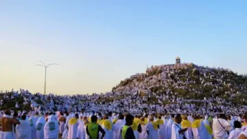 Des milliers de pèlerins en ihram se rassemblent au sommet de la colline de la montagne Arafat pendant le Hajj, sous un ciel bleu. L'image capture l'importance spirituelle de ce moment, avec des drapeaux et des pancartes visibles parmi la foule.