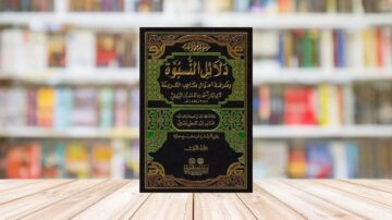 غلاف كتاب دلائل النبوة للبيهقي بتصميم أنيق يتضمن نصوصاً باللون الذهبي، موضوع على طاولة خشبية مع خلفية تضم رفوف كتب