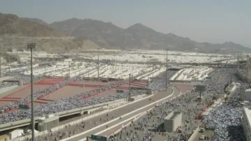 Vue panoramique de la plaine de Mina pendant le Hajj, avec des milliers de pèlerins en blanc s'étendant sur le terrain. Les montagnes en arrière-plan et les infrastructures routières sont visibles, illustrant l'ampleur de cet événement religieux majeur.