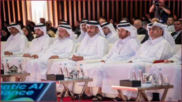 مجموعة من الرجال يرتدون الملابس التقليدية العربية، يجلسون في صفوف خلال حدث رسمي. يركزون على المتحدث، مع وجود جمهور خلفهم. تظهر في المقدمة زجاجات مياه ومواد مكتبية على الطاولات. يعكس المشهد أهمية الفعالية في مجال الذكاء الاصطناعي.
