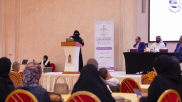 امرأة ترتدي النقاب تتحدث في مؤتمر، بينما يجلس عدد من الرجال والنساء في الصفوف الأمامية. خلفية الحدث تحتوي على لافتة تحمل شعار البنك الدولي الإسلامي، مع شاشة عرض تعرض المحتوى.