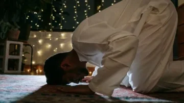 Homme en tenue traditionnelle priant à genoux sur un tapis, dans une ambiance tamisée avec des lumières féeriques en arrière-plan.