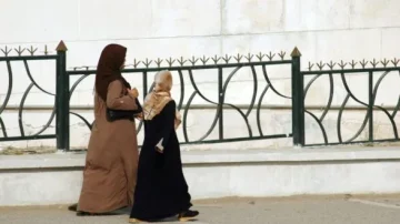 Deux femmes marchent le long d'une clôture. L'une porte une robe marron et un foulard, tandis que l'autre est vêtue de noir et porte un hijab. L'arrière-plan montre un mur clair avec une architecture simple.