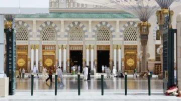 Vue de l'extérieur de la mosquée du Prophète à Médine, montrant des portes ornées et des colonnes. Des fidèles se déplacent dans la cour, avec des ombres projetées par les auvents. L'architecture islamique est mise en valeur par des motifs géométriques et des détails dorés.