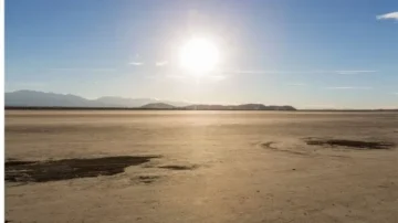 Vue panoramique d'un paysage désertique avec un sol aride et le soleil brillant à l'horizon. Des montagnes se dessinent au loin sous un ciel dégagé, créant une atmosphère sereine et naturelle.