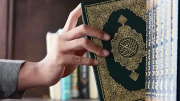 Main dans une bibliothèque, retirant un exemplaire du Coran d'une étagère. Le livre présente une couverture verte ornée de motifs dorés. En arrière-plan, d'autres livres sont visibles, créant une ambiance de sérénité et de culture.