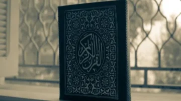 Coran posé sur une surface, avec une couverture ornée de motifs arabesques, devant une fenêtre à barreaux, créant une ambiance sereine et spirituelle.