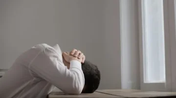 Homme en chemise blanche assis à une table, la tête reposant sur ses bras, dans une ambiance de solitude et de réflexion près d'une fenêtre.