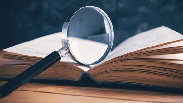 عدسة مكبرة موضوعة على كتاب مفتوح، مع تركيز على النص داخل الكتاب والخلفية غير واضحة، تعكس مفهوم البحث والدراسة.